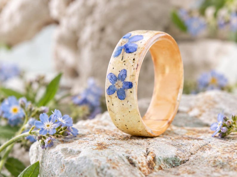 May include: A band ring featuring a smooth, light beige, speckled surface embedded with small, vibrant blue five-petaled flowers. The ring has a polished, rounded exterior and a warm, light-toned interior finish, showcasing a natural aesthetic.