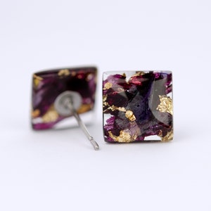 May include: Square stud earrings with a clear resin base and a design of purple and gold flower petals.