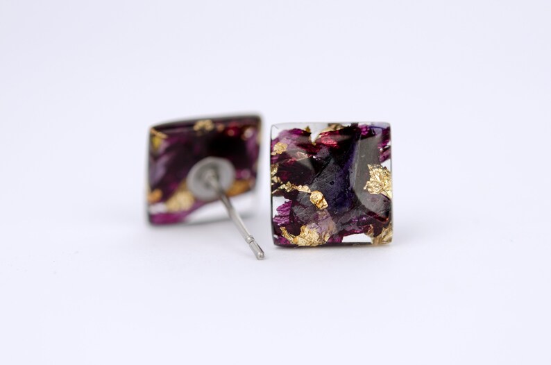 May include: Square stud earrings with a clear resin base and a design of purple and gold flower petals.