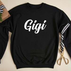 May include: Black sweatshirt with white text that says "Gigi" on the front. The sweatshirt is laying flat on a light brown surface.