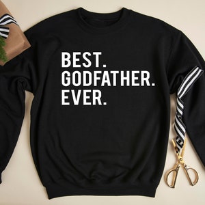 Op de afbeelding: Zwart sweatshirt met de witte tekst "BEST. GODFATHER. EVER.". Een ingepakt cadeau met een zwart-wit gestreept lint en een gouden schaar liggen naast het sweatshirt.