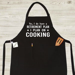 Peut inclure: Tablier noir avec deux poches et des bretelles r&eacute;glables. Le tablier porte une inscription blanche qui dit "Oui, j'ai un PLAN DE RETRAITE * J'AI L'INTENTION DE * CUISINER".