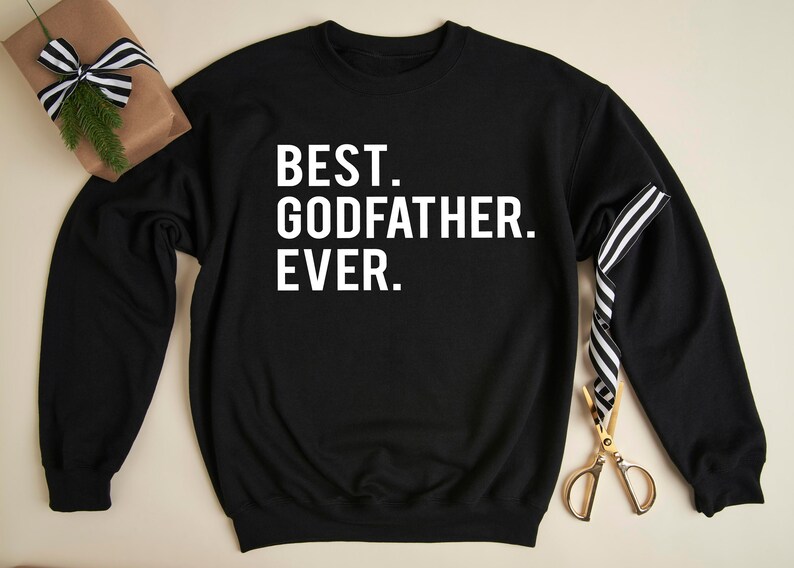 Op de afbeelding: Zwart sweatshirt met de witte tekst "BEST. GODFATHER. EVER.". Een ingepakt cadeau met een zwart-wit gestreept lint en een gouden schaar liggen naast het sweatshirt.