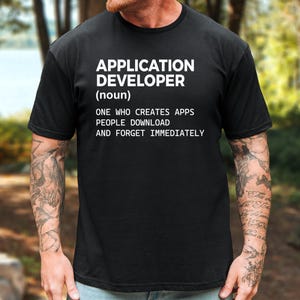 May include: A black short-sleeved t-shirt featuring white text that reads: APPLICATION DEVELOPER (noun) ONE WHO CREATES APPS PEOPLE DOWNLOAD AND FORGET IMMEDIATELY. The shirt has a crew neckline and a standard fit, displaying the printed text clearly.