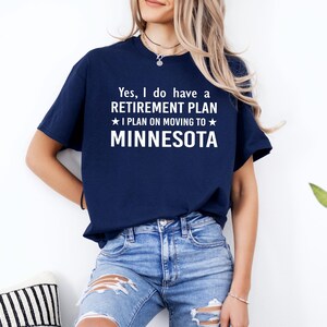 May include: Navy blue t-shirt with white text: "Yes, I do have a RETIREMENT PLAN I PLAN ON MOVING TO MINNESOTA." The shirt is worn by a person in blue jeans. The text is centered on the front of the shirt.