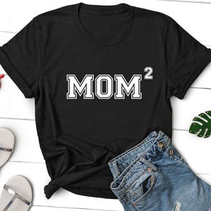 K&ouml;nnte beinhalten: Schwarzes T-Shirt mit wei&szlig;em Text, der "MOM^2" lautet. Das Shirt ist auf einer wei&szlig;en Oberfl&auml;che mit einer Jeans und einem gr&uuml;nen Blatt zu sehen.
