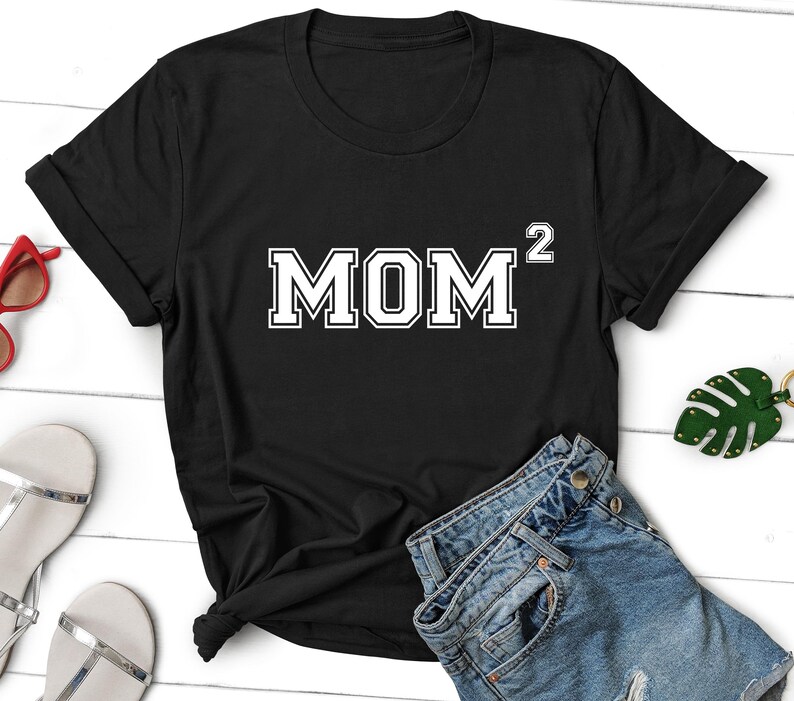 K&ouml;nnte beinhalten: Schwarzes T-Shirt mit wei&szlig;em Text, der "MOM^2" lautet. Das Shirt ist auf einer wei&szlig;en Oberfl&auml;che mit einer Jeans und einem gr&uuml;nen Blatt zu sehen.