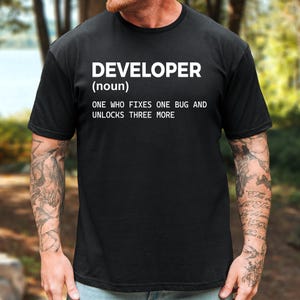 May include: A black short-sleeved t-shirt featuring white text that reads: DEVELOPER (noun) ONE WHO FIXES ONE BUG AND UNLOCKS THREE MORE. The shirt has a crew neckline and a standard fit, displaying the printed definition clearly on the chest area.