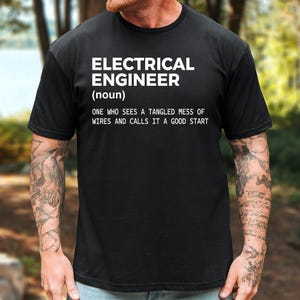 May include: A black short-sleeved t-shirt featuring white text that reads: ELECTRICAL ENGINEER (noun) ONE WHO SEES A TANGLED MESS OF WIRES AND CALLS IT A GOOD START. The shirt has a crew neckline and a standard fit, displaying the printed graphic clearly.