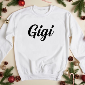 May include: White sweatshirt with the word "Gigi" printed in black cursive lettering. The sweatshirt is surrounded by pine branches, red ornaments, and candy canes.