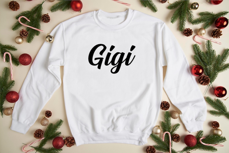 May include: White sweatshirt with the word "Gigi" printed in black cursive lettering. The sweatshirt is surrounded by pine branches, red ornaments, and candy canes.