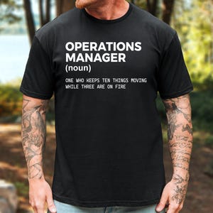 May include: A black short-sleeved t-shirt featuring white text that reads: OPERATIONS MANAGER (noun) ONE WHO KEEPS TEN THINGS MOVING WHILE THREE ARE ON FIRE. The shirt has a crew neckline and a standard fit, displaying the printed graphic on the chest.