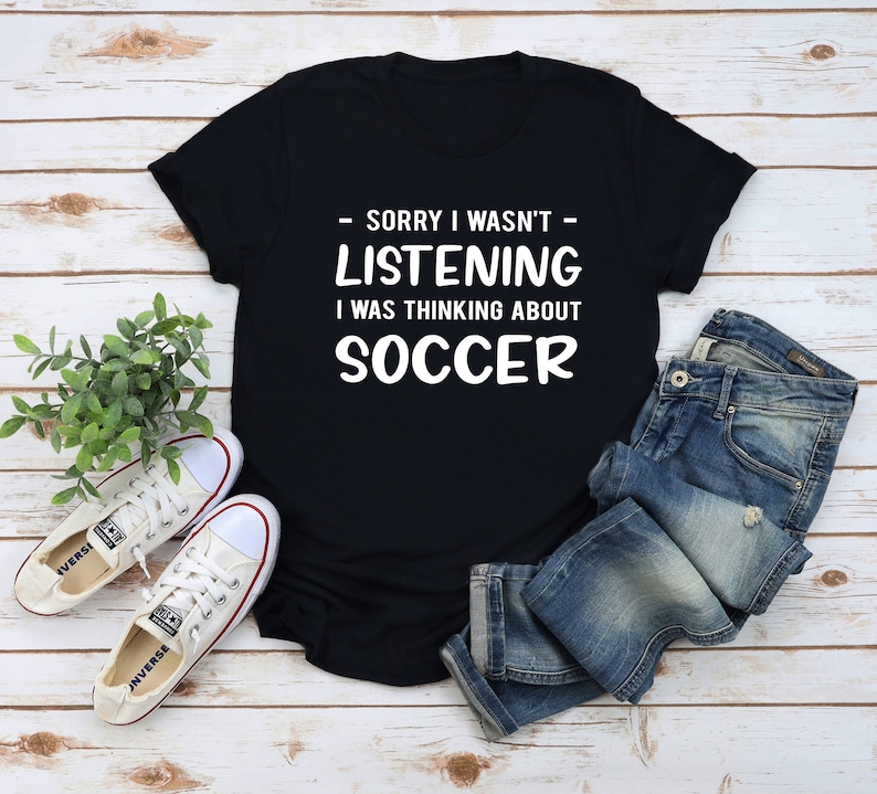 K&ouml;nnte beinhalten: Schwarzes T-Shirt mit wei&szlig;em Text, der "Sorry, ich habe nicht zugeh&ouml;rt, ich habe an Fu&szlig;ball gedacht" lautet. Das Shirt liegt auf einer Holzoberfl&auml;che mit einem Paar wei&szlig;en Sneakers und einer blauen Jeans.