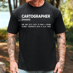 May include: A black short-sleeved t-shirt featuring white text that reads: CARTOGRAPHER (noun) ONE WHO GETS PAID TO MAKE A ROUND PLANET COOPERATE WITH A FLAT PAGE. The shirt has a crew neckline and a standard fit, displaying the printed definition clearly.