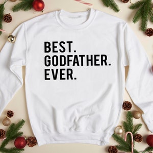 Op de afbeelding: Witte sweatshirt met de zwarte tekst "BEST. GODFATHER. EVER.". De afbeelding is versierd met kerstversieringen, waaronder rode ballen, dennentakken en zuurstokken.