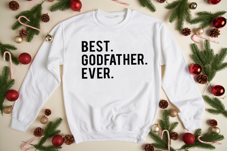 Op de afbeelding: Witte sweatshirt met de zwarte tekst "BEST. GODFATHER. EVER.". De afbeelding is versierd met kerstversieringen, waaronder rode ballen, dennentakken en zuurstokken.
