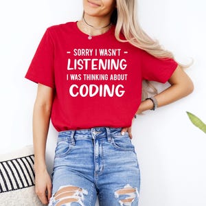 May include: A red t-shirt with the text "- SORRY I WASN'T LISTENING I WAS THINKING ABOUT CODING" in white. The shirt is paired with blue jeans. The overall theme is related to coding and computer science.