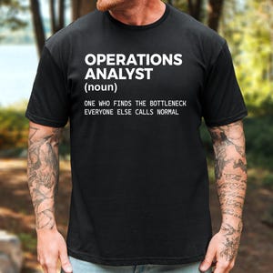 May include: A black short-sleeved t-shirt featuring white text that reads: OPERATIONS ANALYST (noun) ONE WHO FINDS THE BOTTLENECK EVERYONE ELSE CALLS NORMAL. The shirt has a crew neckline and a standard fit, displaying the printed graphic on the chest.