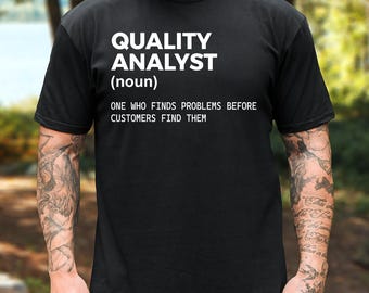 May include: A black short-sleeved t-shirt featuring white text that reads: QUALITY ANALYST (noun) ONE WHO FINDS PROBLEMS BEFORE CUSTOMERS FIND THEM. The shirt has a crew neckline and a standard fit, displaying the printed definition clearly on the chest.