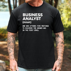 May include: A black short-sleeved t-shirt featuring white text that reads: BUSINESS ANALYST (noun) ONE WHO ATTENDS FIVE MEETINGS TO DISCOVER THE ANSWER WAS IN THE FIRST EMAIL. The shirt has a crew neckline and a standard fit, displaying clear white lettering.