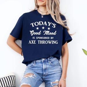 May include: Navy blue t-shirt with the text "TODAY'S Good Mood is SPONSORED BY AXE THROWING" in white. The shirt has a crew neck and short sleeves. The model is wearing blue jeans. A plant is visible in the background.