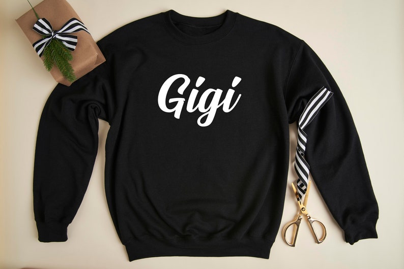 May include: Black sweatshirt with white text that says "Gigi" on the front. The sweatshirt is laying flat on a light brown surface.