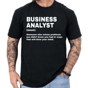 May include: Black t-shirt with the words "BUSINESS ANALYST" in large white letters. Below, the definition of a business analyst is printed in white. The shirt is a crew neck style.