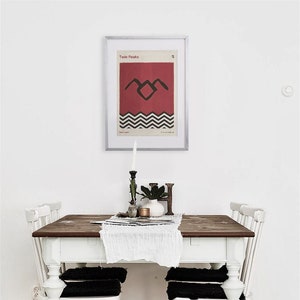 May include: A white dining table with four white chairs and black cushions. The table has a wooden top and is set with a white tablecloth and a framed poster of the Twin Peaks logo.