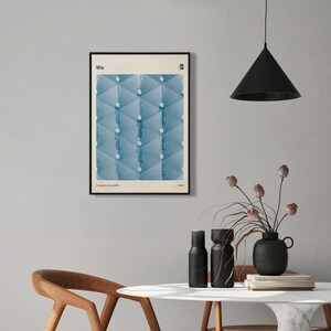 May include: A framed poster with a geometric design in shades of blue. The poster has the word "We" at the top and the text "Yevgeny Zamlyatin, 1921" at the bottom. The poster is hanging on a light gray wall above a white marble table with two wooden chairs.