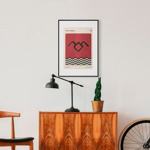 May include: A framed poster with a minimalist design featuring the iconic black and white "Twin Peaks" logo on a red background. The poster is hanging on a white wall above a wooden cabinet.