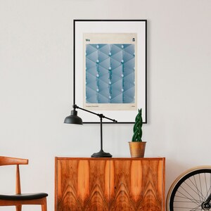 May include: A framed poster with a blue geometric design and the word "We" in white text. The poster is hanging on a white wall above a wooden cabinet with two doors. A black lamp with a white shade is on the cabinet, and a potted plant is to the right of the lamp. A bicycle wheel is on the right side of the image.