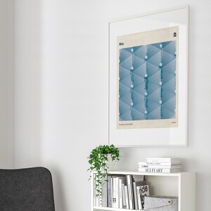 May include: A white bookshelf with four shelves filled with books and a glass vase. The bookshelf is against a white wall with a framed print of a geometric design in blue and white. The print has the text "We" and "Evgeny Zamjatin 1924".