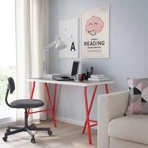 K&ouml;nnte beinhalten: Ein modernes Heimb&uuml;ro mit einem wei&szlig;en Schreibtisch mit roten Beinen, einem grauen Drehstuhl und einer wei&szlig;en, verstellbaren Schreibtischlampe. Ein Laptop, B&uuml;cher und eine Tasse stehen auf dem Schreibtisch. Zwei gerahmte Poster h&auml;ngen an der Wand.