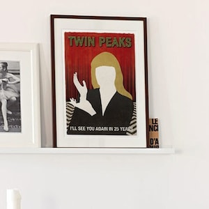 Op de afbeelding: Een zwarte rieten fauteuil met een witte nep bont deken en een zwart kussen. Een kleine grijze tafel met een kaars en boeken staat naast de stoel. Twee ingelijste foto's hangen aan de muur achter de stoel. De foto links toont een ballerina in een zwarte maillot en tutu. De foto rechts is een poster met de tekst "Twin Peaks" en een silhouet van een vrouw in een rode jurk.