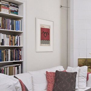 May include: A white couch with red and brown throw pillows in a living room with a white tiled fireplace and a bookshelf filled with books. A framed poster with the text "Twin Peaks" and a black and white graphic of a bird with a heart in its chest is hanging on the wall.