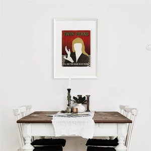 Op de afbeelding: Een witte eettafel met een donkerhouten blad en vier witte stoelen. De stoelen hebben zwarte kussens. Een ingelijste poster met de tekst "Twin Peaks" en een silhouet van een vrouw hangt aan de muur boven de tafel.