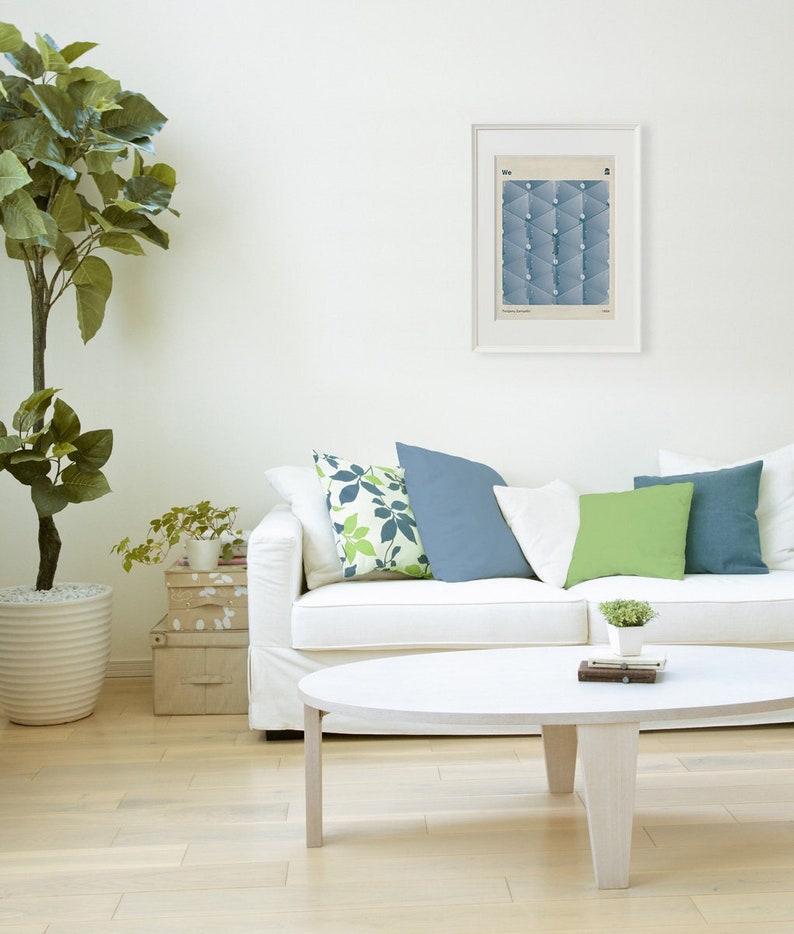 May include: A white couch with green, blue, and white patterned throw pillows. A small potted plant sits on a coffee table in front of the couch. The coffee table is oval shaped and white. The couch is in a room with white walls and a framed print on the wall.
