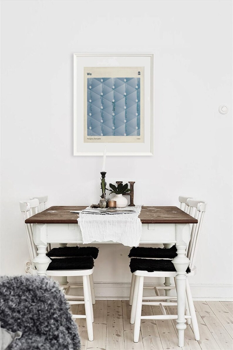 May include: A white farmhouse style dining table with a wood top and four white chairs. The chairs have black cushions. A white tablecloth with a lace trim is on the table. A framed print with a blue geometric design and the word "We" is hanging on the wall above the table.