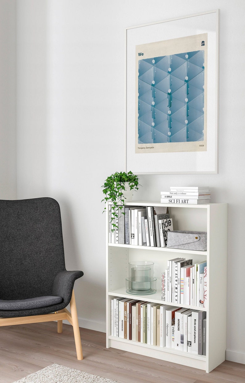 May include: A white bookshelf with four shelves filled with books and a glass vase. The bookshelf is against a white wall with a framed print of a geometric design in blue and white. The print has the text "We" and "Evgeny Zamjatin 1924".