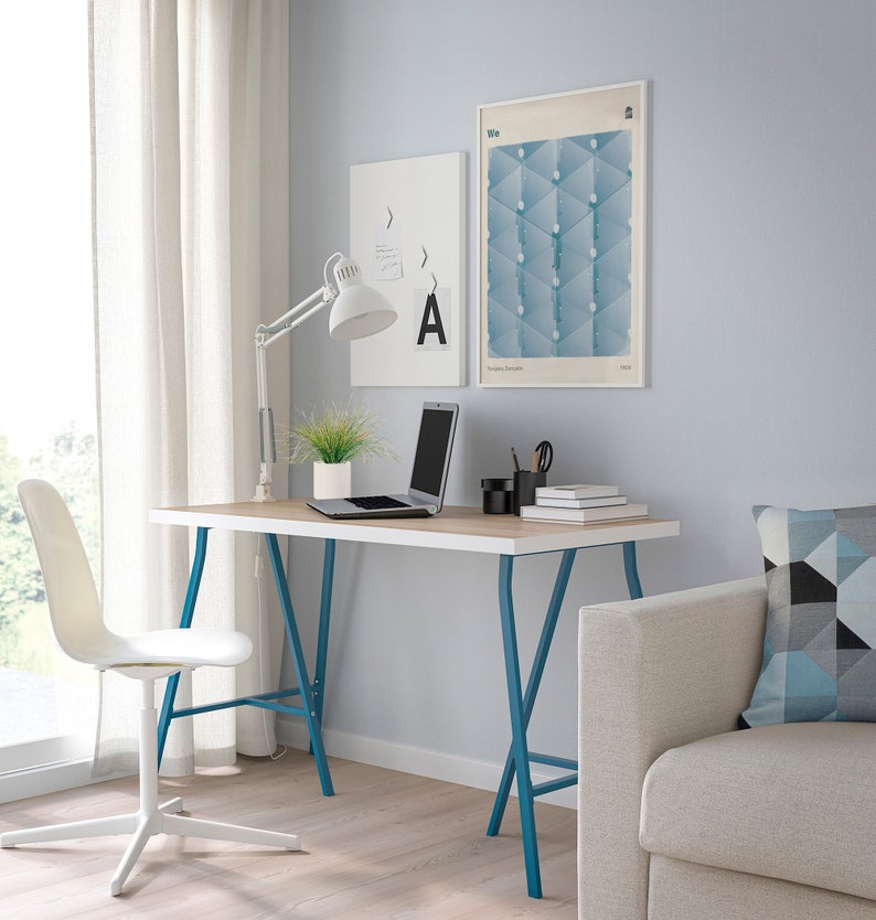 May include: A white desk with a light wood top and blue metal legs. A laptop, a plant, and a lamp are on the desk. A white chair is in front of the desk. There are two framed prints on the wall behind the desk. The print on the left is white with a black arrow and the letter A. The print on the right is blue and white with the word "We" at the top.