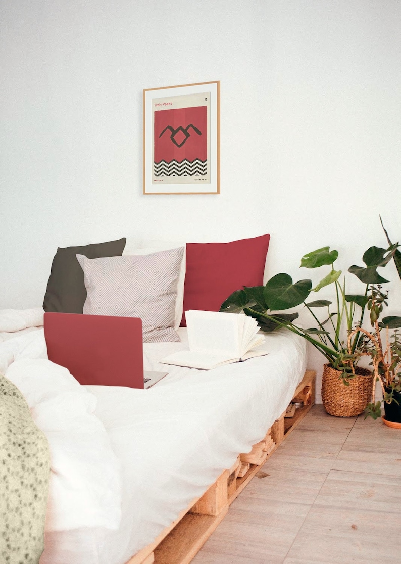 May include: A white bed with a red laptop, a white book, and a red, gray, and white patterned pillow. The bed is made of wooden pallets and has a white blanket. There is a framed poster on the wall behind the bed with the text "Twin Peaks" and a black and white graphic of a bird. There are two green plants in pots on the floor next to the bed.