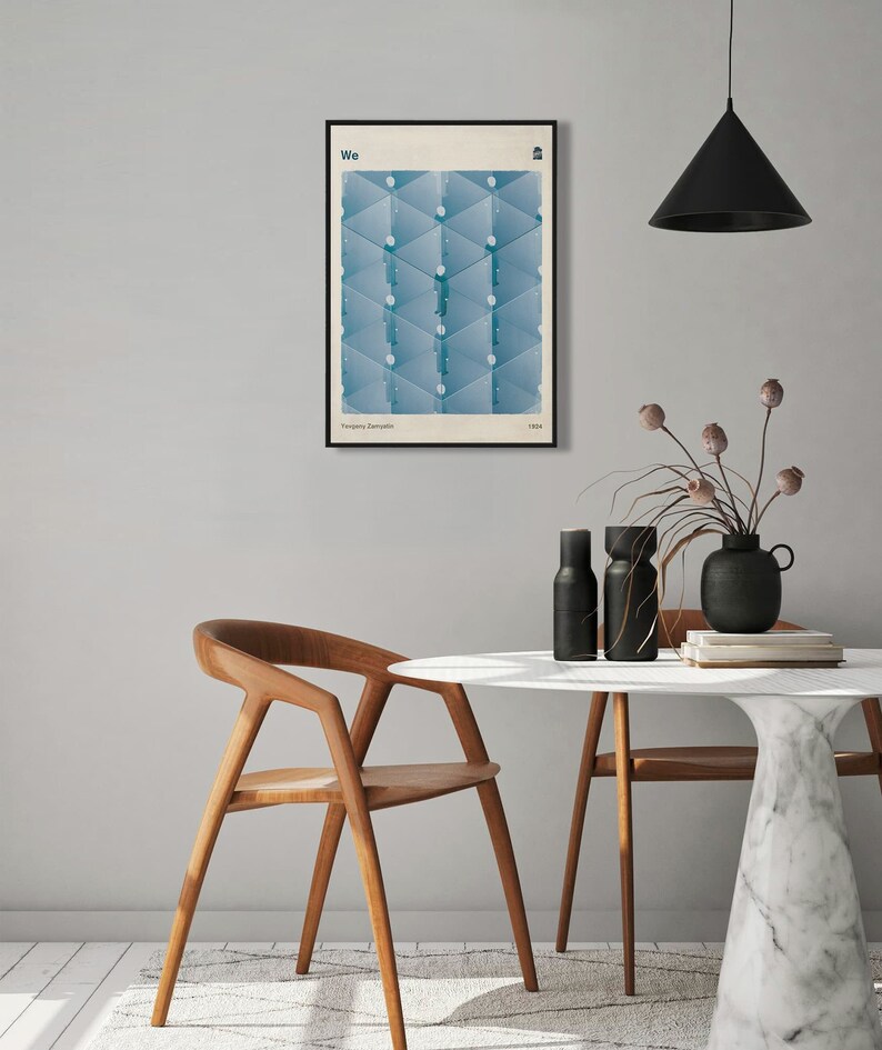 May include: A framed poster with a geometric design in shades of blue. The poster has the word "We" at the top and the text "Yevgeny Zamlyatin, 1921" at the bottom. The poster is hanging on a light gray wall above a white marble table with two wooden chairs.