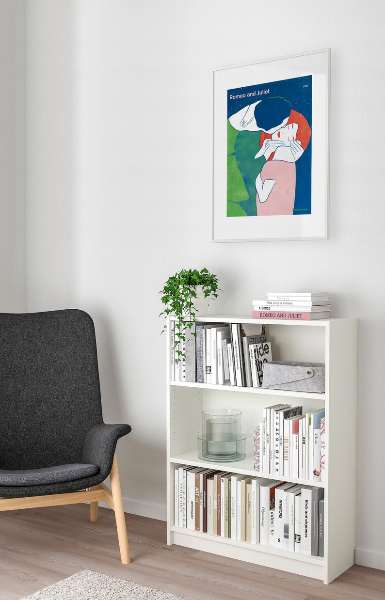 May include: A white bookshelf with three shelves filled with books and decorative items. The bookshelf is against a white wall with a framed poster of Romeo and Juliet above it. A gray armchair with wooden legs is in the foreground.
