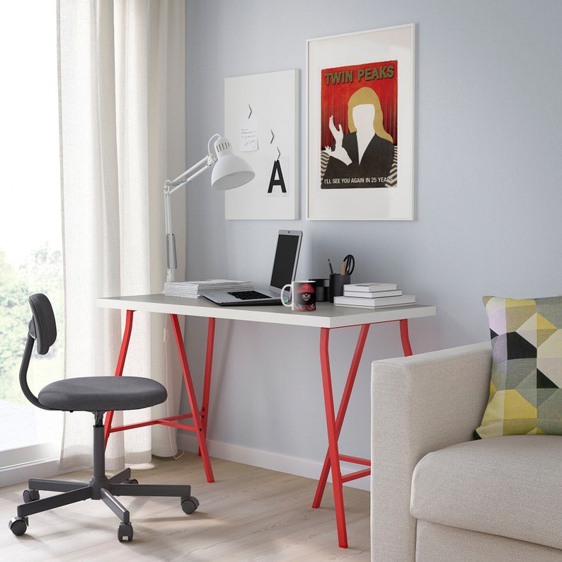 Op de afbeelding: Een wit bureau met rode metalen poten, een laptop, een mok en een stapel boeken. Een grijze bureaustoel met wielen staat op de voorgrond. Op de achtergrond is een witte bank met een geometrisch patroon kussen. Twee posters hangen aan de muur, een met de tekst "Twin Peaks" en de andere met de letter "A".