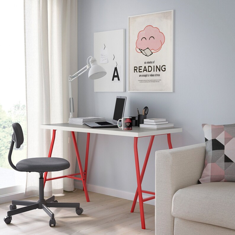 K&ouml;nnte beinhalten: Ein modernes Heimb&uuml;ro mit einem wei&szlig;en Schreibtisch mit roten Beinen, einem grauen Drehstuhl und einer wei&szlig;en, verstellbaren Schreibtischlampe. Ein Laptop, B&uuml;cher und eine Tasse stehen auf dem Schreibtisch. Zwei gerahmte Poster h&auml;ngen an der Wand.