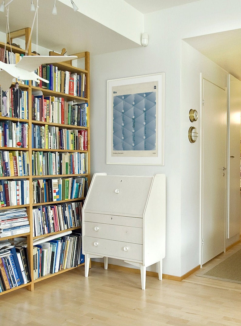 May include: A white wooden secretary desk with three drawers and a drop-down front. The desk is in a room with a bookshelf filled with books and a framed poster with the word "We" on it.