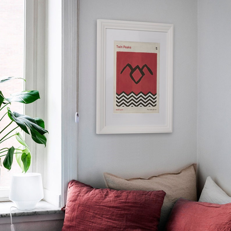 May include: A white framed poster with the text "Twin Peaks" and a black and red graphic of a stylized mountain range with the text "Fire walk with me". The poster is hanging on a white wall above a red and beige couch.