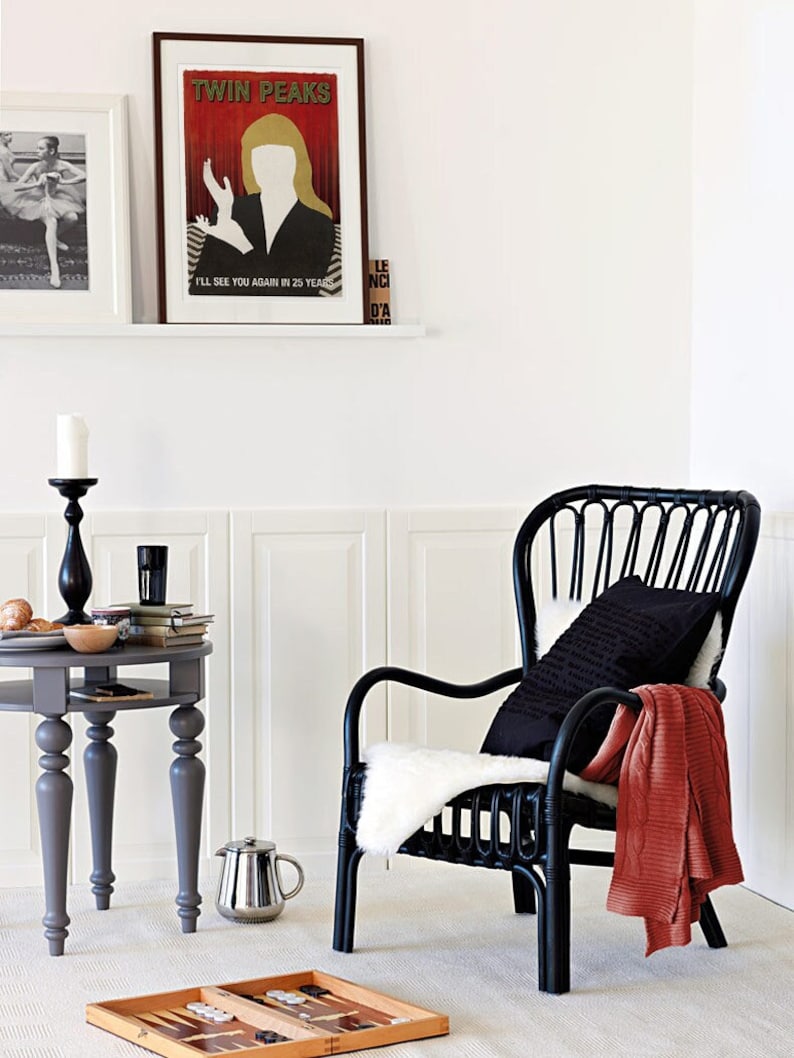 Op de afbeelding: Een zwarte rieten fauteuil met een witte nep bont deken en een zwart kussen. Een kleine grijze tafel met een kaars en boeken staat naast de stoel. Twee ingelijste foto's hangen aan de muur achter de stoel. De foto links toont een ballerina in een zwarte maillot en tutu. De foto rechts is een poster met de tekst "Twin Peaks" en een silhouet van een vrouw in een rode jurk.