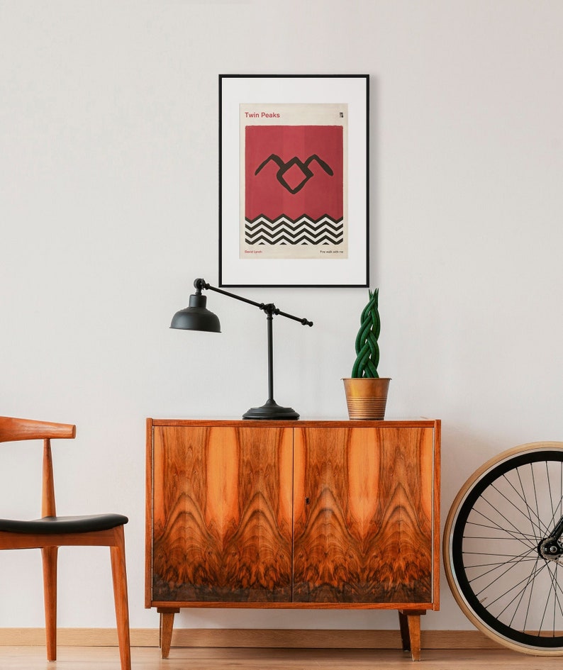 May include: A framed poster with a minimalist design featuring the iconic black and white "Twin Peaks" logo on a red background. The poster is hanging on a white wall above a wooden cabinet.