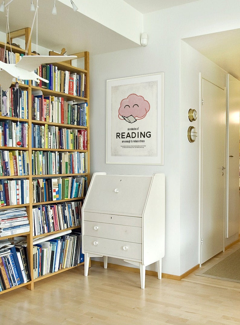 K&ouml;nnte beinhalten: Innenraum mit einem B&uuml;cherregal aus Holz voller B&uuml;cher, einem wei&szlig;en Schreibtisch mit Schubladen und einem gerahmten Poster mit dem Text "six minutes of READING are enough to reduce stress". Der Raum hat hellfarbene Holzb&ouml;den.